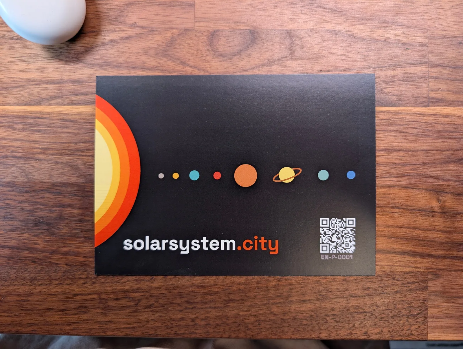 The World Trade Center mission postcard on a wooden table: the sun's layered orange arcs at the left edge, the eight planets in a row across a black field, and the walk's QR code at the lower right.