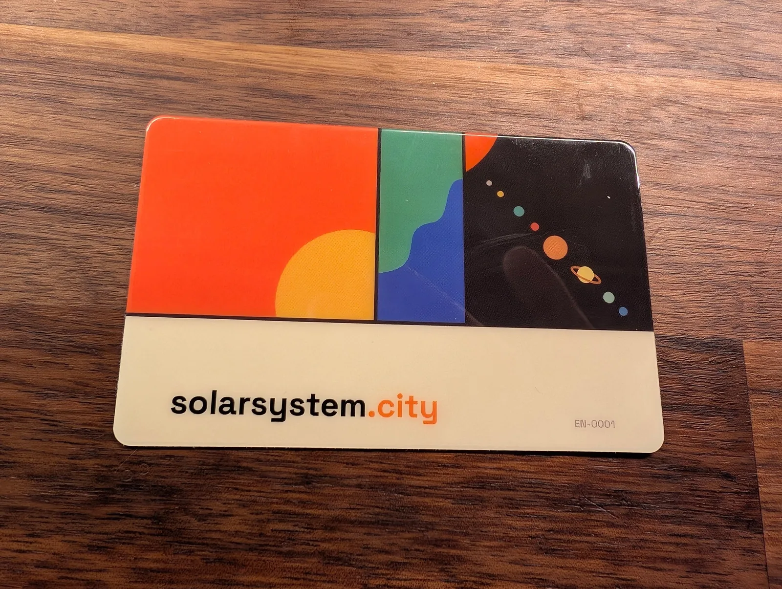 The SolarSystem.city Sun Card on a wooden table: a wallet-sized card printed with a large orange sun, a green-and-blue Earth panel, and the planets in miniature against a black field.
